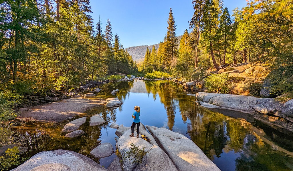 A woman standing on a boulder in the middle of a river.