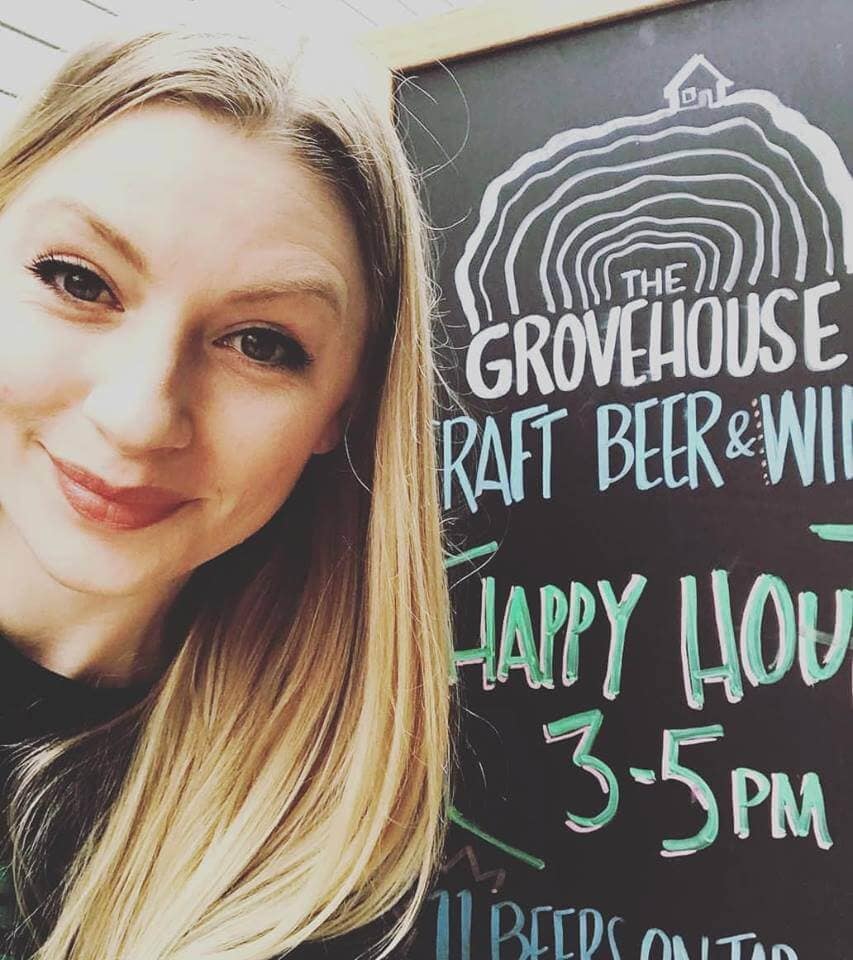 Smiling woman with the Grove House sign showcasing happy hour times