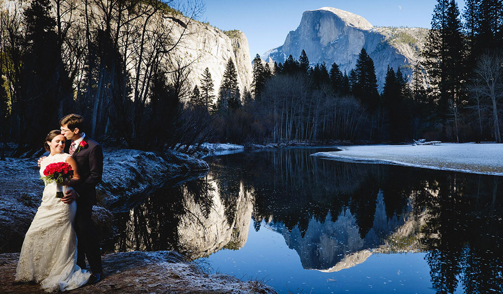 Wedding couple standing by the Merced River in winter with a snowy Half Dome behind.