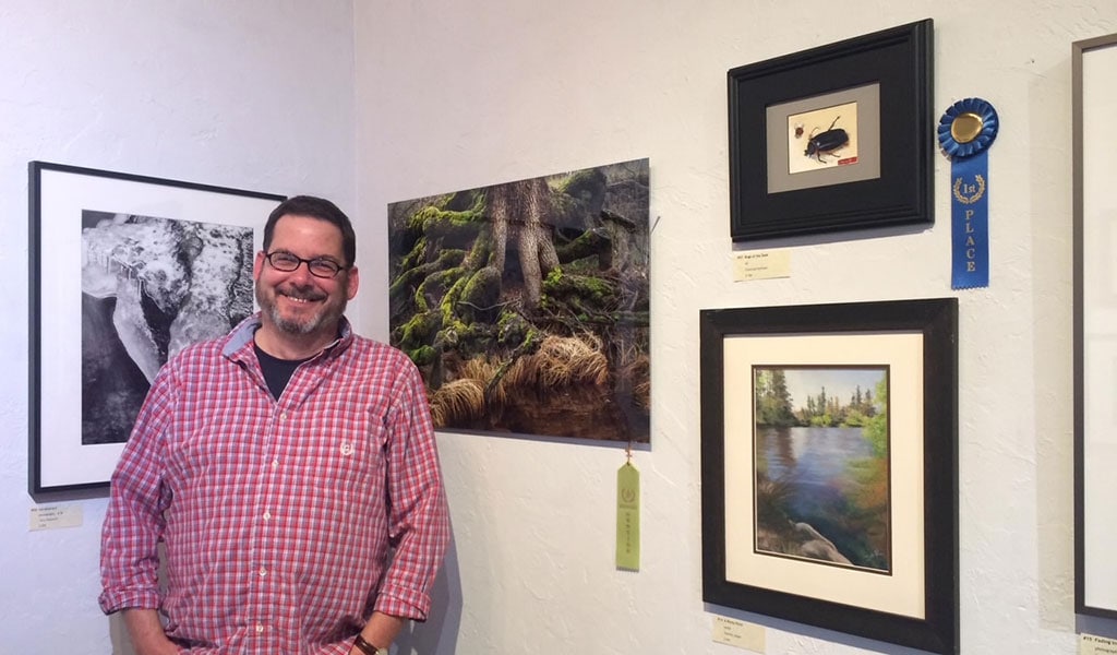 Smiling man standing next to photographs and paintings in an art exhibit.