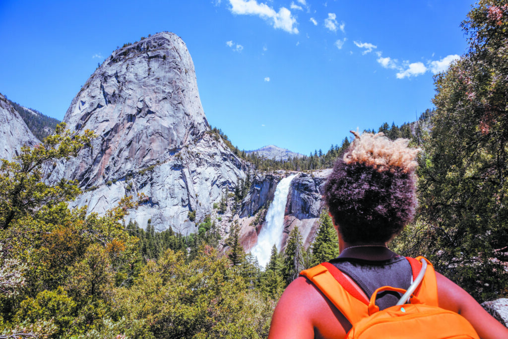 A hiker with curly hair and an orange backpack is photographed from behind, looking out over a dramatic vista of a mountain, forest, and waterfall. In the center, a powerful waterfall cascades over a cliff into a tree-filled valley. To the left, a massive, smooth, rounded granite dome dominates the scene. The sky above is a bright, clear blue with a few small white clouds.
