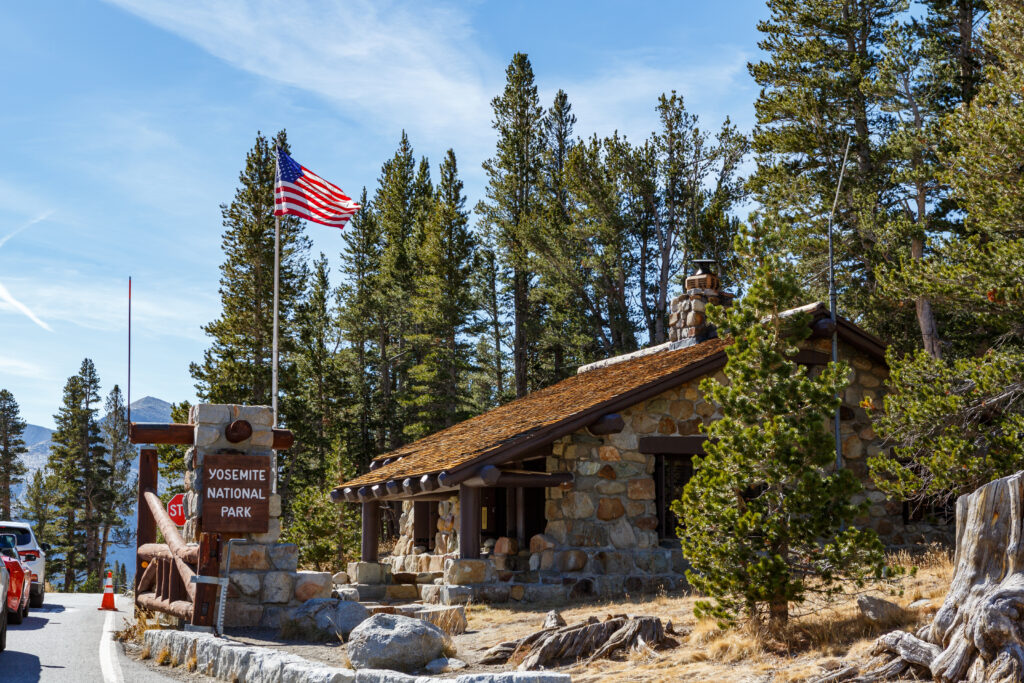 
A rustic, low-slung stone and log building with a dark, shingled roof and a stone chimney stands among tall evergreen trees under a blue sky. To the left, a wooden and stone sign marks the entrance to Yosemite National Park, with a large American flag waving from a tall flagpole behind it. The entrance is near a paved road with orange traffic cones and a vehicle partially visible in the bottom left.