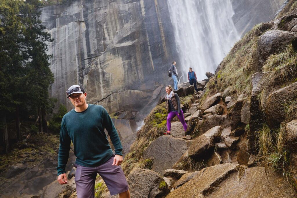 
A low-angle shot on the Mist Trail in Yosemite National Park shows a man in a dark green sweater, gray shorts, a white baseball cap, and sunglasses standing in the left foreground, looking directly at the camera. Behind him, a line of three other hikers is carefully making their way up a steep, rocky path on the right. A woman in bright purple leggings is in the center, ascending the large boulders. The backdrop is dominated by a sheer granite cliff face on the left and the powerful rush of a large waterfall on the right, creating a misty atmosphere.