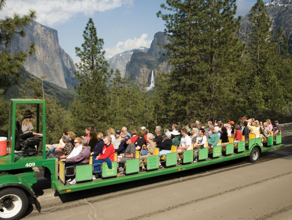 Yosemite Valley Floor Tour. The passengers are seated and facing right, with many looking toward the spectacular natural scenery. In the background, tall evergreen trees line the road, and the iconic granite cliffs of El Capitan and Half Dome are visible, along with a flowing waterfall in the distance. The sky is blue with a few white clouds.